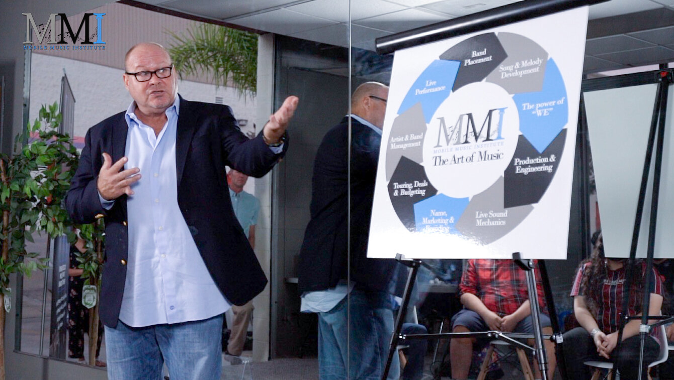 A speaker gestures toward a chart explaining MMI's curriculum at a music sponsorship presentation.