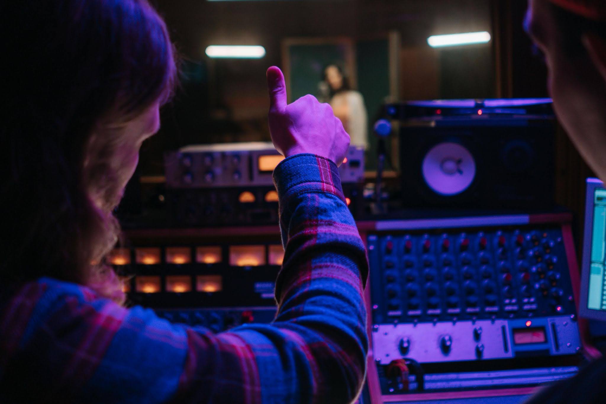 Studio producer seated at a mixing console, giving a thumbs up to a vocalist recording behind the glass.